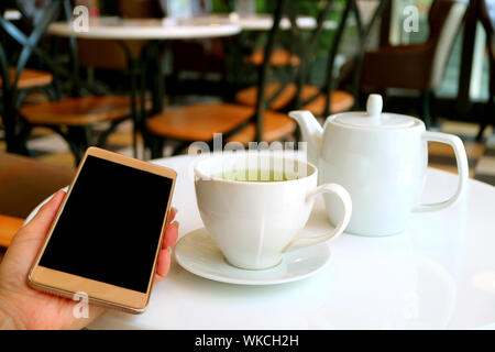 One Cup of Hot Green Tea and Teapot on Cafe Table with Female Hand Holding a Mobile Phone Stock Photo