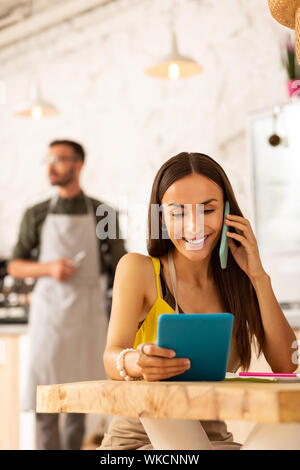 Calling clients. Dark-haired beaming wife calling clients while helping husband with business Stock Photo