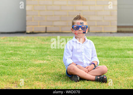 Smiling Australian boy with sunglasses sitting on lawn during Australia Day celebration Stock Photo