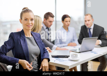 business, technology and office concept - smiling businesswoman with eyeglasses in office with team on the back Stock Photo