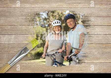 Composite image of senior couple on bikes in the park against wooden surface with paintbrushes Stock Photo