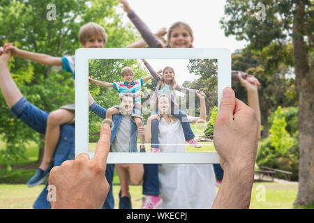 Hand holding tablet pc showing parents carrying kids on shoulders at park Stock Photo