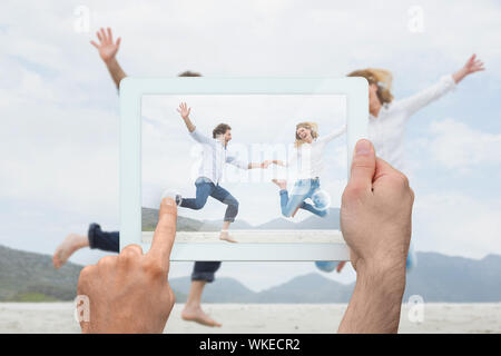 Hand holding tablet pc showing cheerful couple holding hands and jumping at beach Stock Photo