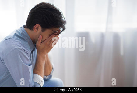 Frustrated Lonely Man Covering Face With Hands Sitting Against Window Stock Photo