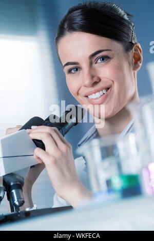 Female researcher using microscope and smiling at camera with beakers on foreground. Stock Photo