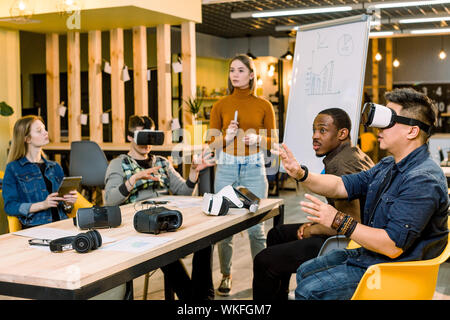 Young woman leader of the project speaking to her colleagues standing near the whote office board. Multiracial team of people having fun wearing Stock Photo