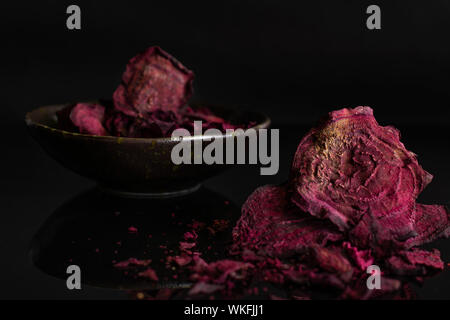 Lot of slices of dried red beetroot few are in the front and the rest is in dark ceramic bowl isolated on black glass Stock Photo