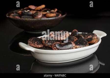 Lot of whole stained purple bean in white oval ceramic bowl in dark ceramic bowl isolated on black glass Stock Photo