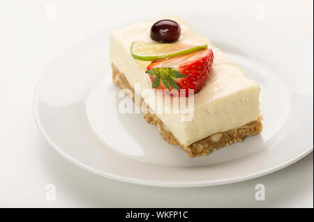 Slice of delicious tangy lemon cheesecake on a biscuit base topped with fresh strawberry and a slice of lemon or lime Stock Photo