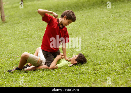 Two young brothers fighting and hitting on grass in park, with older boy sitting over the younger Stock Photo