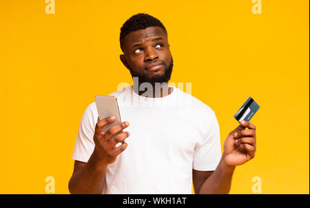 Doubtful african guy holding cellphone and credit card Stock Photo