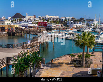 Marina Rubicon harbour resort Playa Blanca clear blue sky sun warm overview, single lady strolling on boardwalk leading to restaurants and bars with luxury sea inlet marina moorings landscape vista view Marina Rubicon Playa Blanca Lanzarote Canary Islands Spain Stock Photo