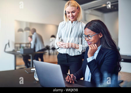 Two smiling diverse businesswomen looking at something on a laptop while working together at an office desk Stock Photo