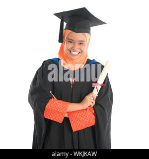 Portrait of smiling Asian female Muslim student in graduate gown showing graduation diploma standing isolated on white background. Stock Photo