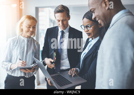Diverse group of smiling businesspeople standing in a bright modern office discussing work together and using a laptop Stock Photo