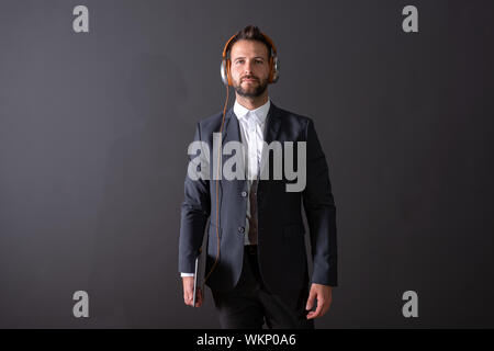 A serious handsome young businessman in a suit holding a tablet and listening to music in front of a dark grey background in a studio. Stock Photo