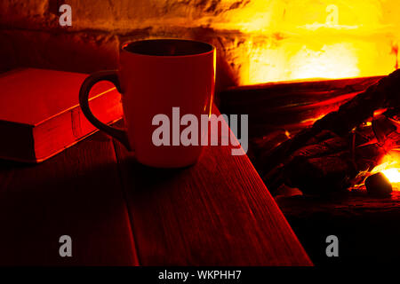A cup of coffee or tea and an old book on a wooden table near the fireplace in a dark room. The concept of rest Stock Photo