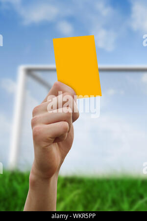 Hand holding up yellow card against goalpost on grass under blue sky Stock Photo