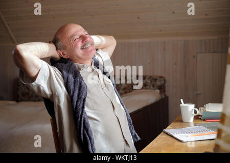 Relaxed senior hispanic man holding hands behind head and smiling sitting at the table. Happy day dreamer concept Stock Photo