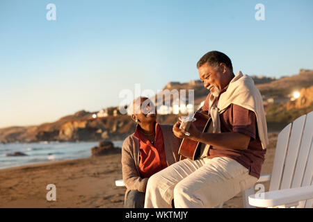 Smiling senior woman listening to her husband play an acoustic guitar on a beach. Stock Photo