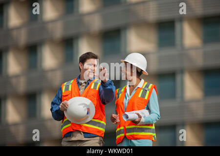Two engineers in high visibility vests and hardhats discuss building plans on a construction site. Stock Photo