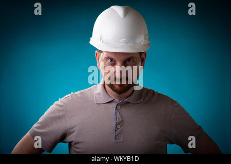 Construction worker with beard and white safety helmet looking stern into the camera over protective glasses against a studio background and blue spot Stock Photo
