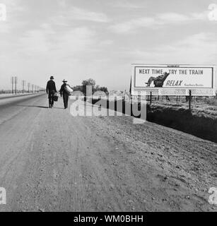 Rear View of Two Men Walking toward Los Angeles, California, USA, on Dirt Road near Billboard that says 'Next Time Try the Train. Relax.', Dorothea Lange, U.S. Farm Security Administration, March 1937 Stock Photo