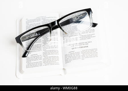 An open white portable pocket bible with a pair of clear reading glasses set on plain white background. Stock Photo