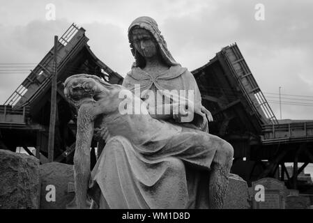 A tombstone depicting Mary holding Jesus in front of the partially demolished  Kosciuszko bridge in Calvary Cemetery in Maspeth, Queens, New York. Stock Photo