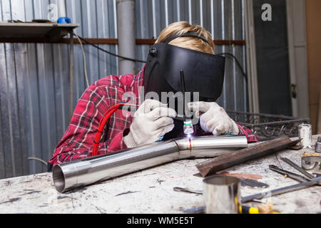 Young guy welder in a checkered red shirt welds a stainless steel pipe using agronomic welding to protect his eyes with a mask in an iron workshop. Mo Stock Photo