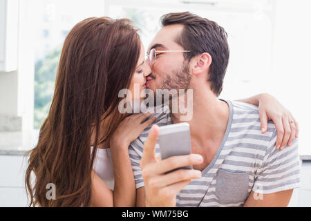 Young man using smartphone while girlfriend kisses him at home in the kitchen Stock Photo