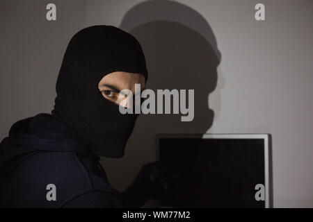 Hacker in balaclava hacking laptop while looking at camera in house Stock Photo