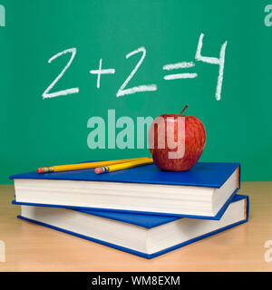 2 + 2 = 4 written on a chalkboard. Books, pencils and an apple on the foreground. Stock Photo