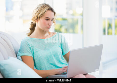 Focused blonde sitting on couch using laptop in bright living room Stock Photo