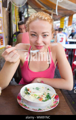 Young carefree adult female traveler eating traditional thai Tom Yum soup from the street stall vendor on Khao San road, Bangkok, Thailand.