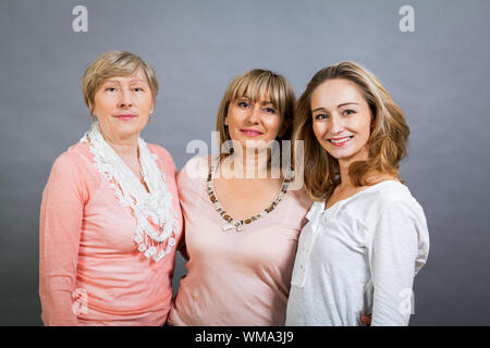 Three generations of attractive women with blond hair and a striking family resemblance posing together arm in arm looking at the camera with friendly Stock Photo