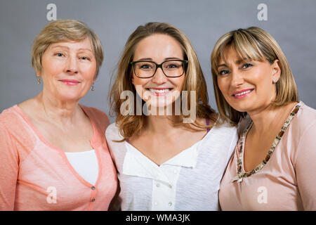 Three generations of attractive women with blond hair and a striking family resemblance posing together arm in arm looking at the camera with friendly Stock Photo