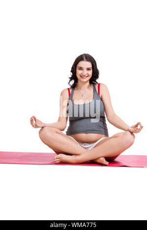Attractive young pregnant woman with a beautiful smile sitting in the lotus position practising yoga and meditating on a gym mat isolated against a wh Stock Photo