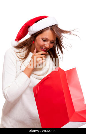 Happy vivacious Christmas shopper wearing a red Santa hat holding up a colorful red shopping bag with a beautiful beaming smile, isolated on white wit Stock Photo