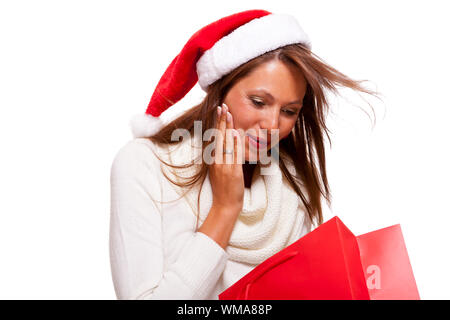 Happy vivacious Christmas shopper wearing a red Santa hat holding up a colorful red shopping bag with a beautiful beaming smile, isolated on white wit Stock Photo