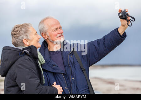 Elderly couple taking a self portrait on a compact digital camera posing in the open air and sunshine with their heads close together smiling at the l Stock Photo