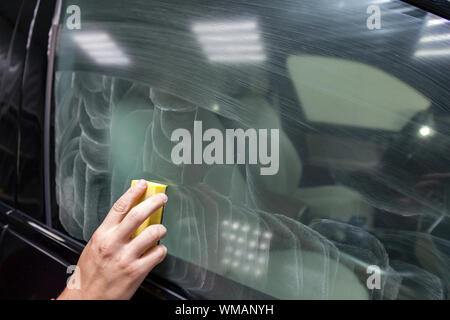 The man’s hand applies a degreasing cleaner with a yellow sponge to the side window of the car to apply a protective nano ceramic coating. Auto servic Stock Photo