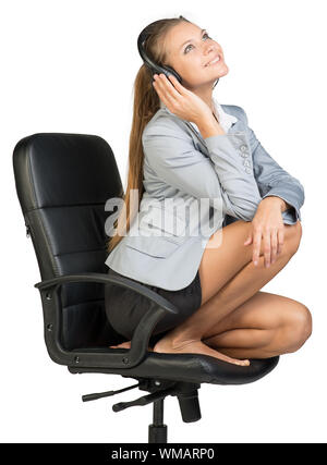 Businesswoman in headset sitting on office chair with legs, looking upwards, smiling. Isolated over white background Stock Photo