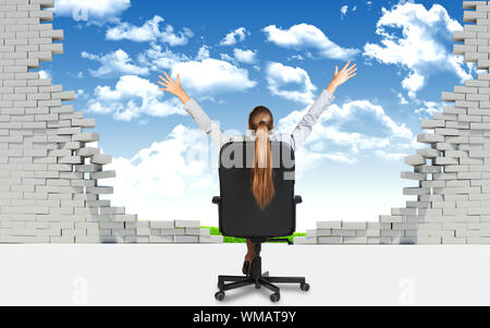 Businesswoman sitting on the office chair with her hands outstretched, in front of broken wall with blus sky and green field behind it. Stock Photo