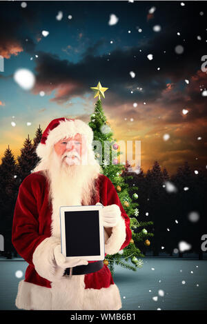 Santa presents a tablet PC against christmas tree in snowy landscape Stock Photo