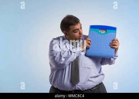 Obese man in formal clothes holding weighing scale with both hands and looking at it Stock Photo