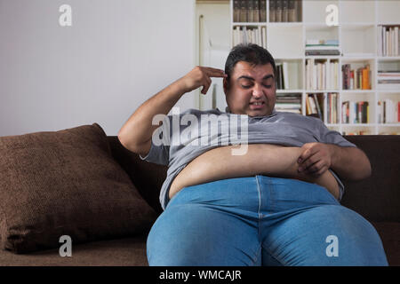Disgusted obese man sitting on sofa at home holding his fat belly and pointing his fingers to his head Stock Photo