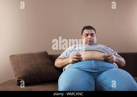 Unhappy obese man sitting on sofa holding his belly fat with both hands Stock Photo