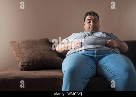 Obese man sitting on sofa holding his belly fat with both hands and looking sad Stock Photo