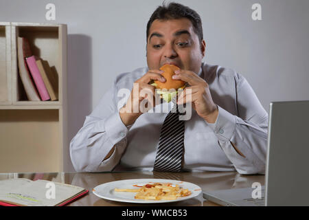 Overweight man eating burger sitting at his desk in office with laptop in front Stock Photo